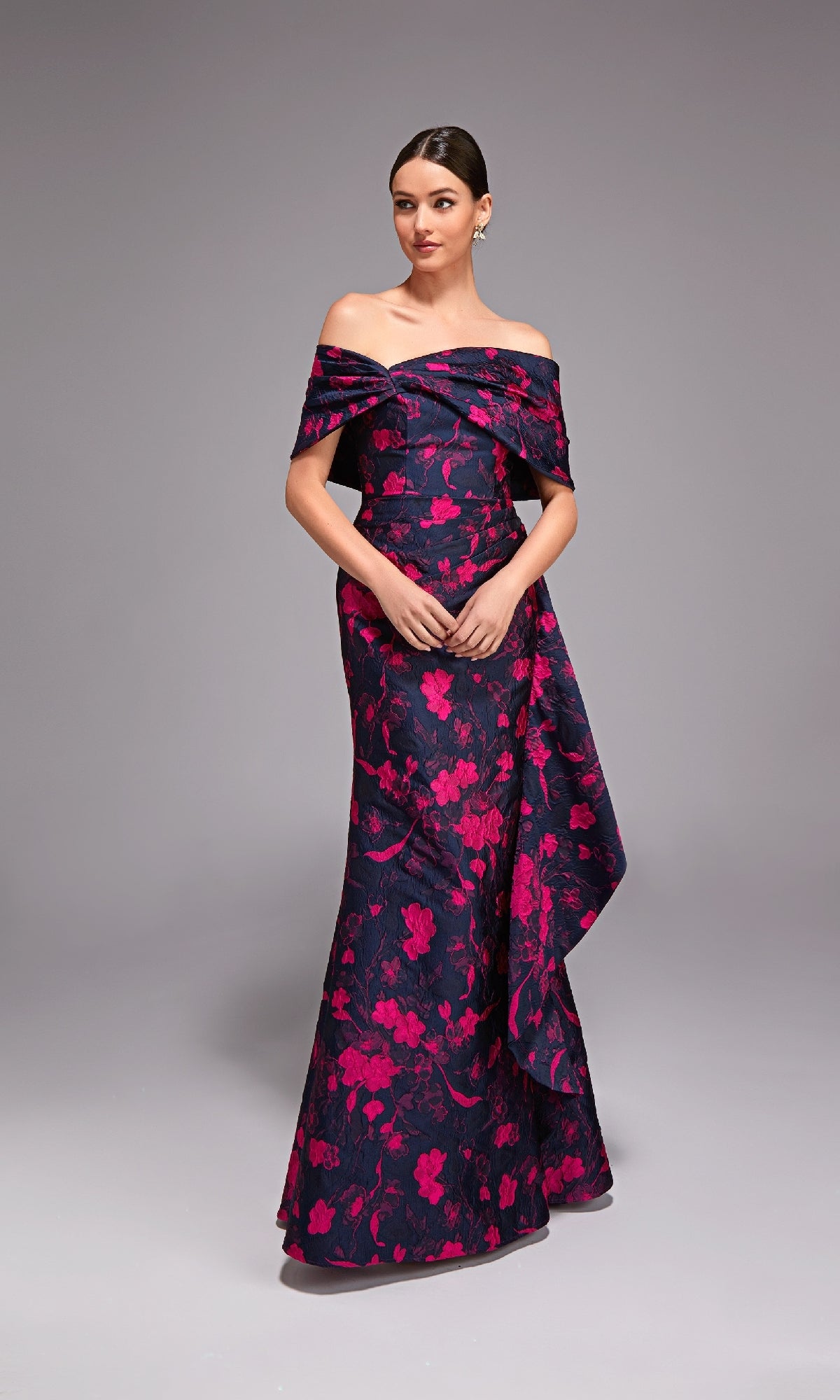 A woman poses against a plain gray backdrop wearing the Alyce 27832 long formal dress in navy/fuchsia, featuring an off-the-shoulder neckline, floor-length design, bright pink floral patterns, and draped floral jacquard fabric.